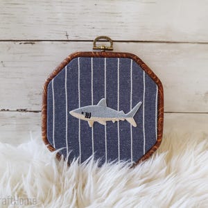 May include: An octagonal embroidery hoop frame featuring a blue fabric background with vertical white pinstripes. Centered on the fabric is an embroidered gray and white shark applique. The frame has a dark brown wood-grain finish and a metal hanger.