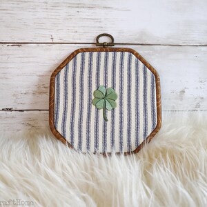 May include: An octagonal embroidery hoop frame containing fabric with a blue and white vertical striped pattern. A green four-leaf clover is embroidered in the center of the fabric. The frame has a dark wood finish and a small metal hanging loop.