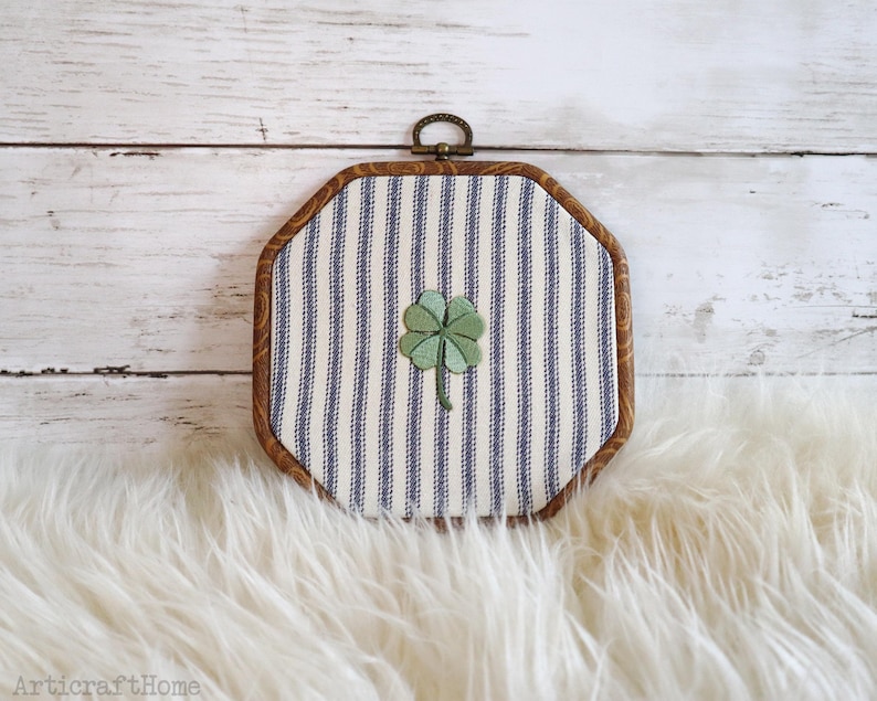May include: An octagonal embroidery hoop frame containing fabric with a blue and white vertical striped pattern. A green four-leaf clover is embroidered in the center of the fabric. The frame has a dark wood finish and a small metal hanging loop.