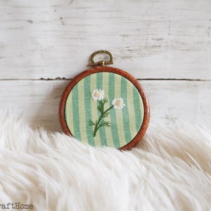 May include: A circular wooden embroidery hoop frame containing light green and white striped fabric. The fabric features a hand-stitched embroidery design of two white flowers with yellow centers and green stems. A small brass hanging loop is at the top.