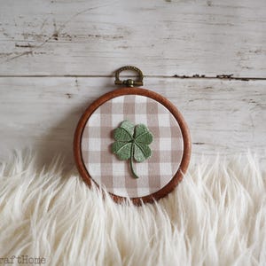 May include: A round, wooden embroidery hoop featuring a green four-leaf clover design. The clover is centered on a tan and white gingham fabric. The hoop has a metal loop at the top for hanging. The background is a white, textured surface.