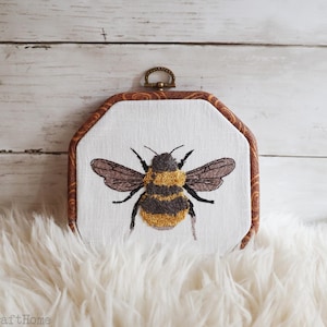 May include: An octagonal embroidery hoop frame containing white fabric with a detailed stitched bumblebee. The bee features textured yellow and dark gray thread, with delicate gray wings. The frame has a brown wood-grain pattern and a metal hanger.