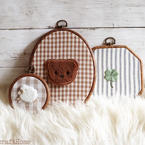 May include: Three embroidery hoops featuring fabric designs: a small round hoop with a white sheep, a large oval hoop with a brown teddy bear face, and a square-cornered hoop with a green four-leaf clover. Each hoop has a dark wood frame.