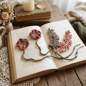 May include: An open book featuring crocheted flower and leaf bookmarks in shades of red, pink, and green. The bookmarks have long, braided stems. The book rests on a wooden surface, with a cup of coffee and a small vase of dried flowers.