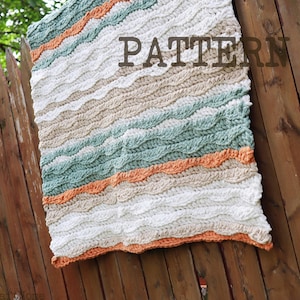 Op de afbeelding: Een gehaakte deken met een golvend, gestructureerd steekpatroon in horizontale strepen van wit, beige, saliegroen en oranje. Het woord PATTERN is in donkerbruine letters gedrukt op het rechterbovenste gedeelte van het textiel.
