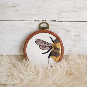 May include: A circular wooden embroidery hoop containing white fabric with a stitched bee design. The bee features textured yellow and grey thread for the body and dark brown thread for the wings. A small bronze-colored metal hanger is attached to the top.