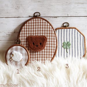 May include: Three embroidery hoops with fabric backgrounds and embroidered motifs. Hoop 1 is small and round with a white sheep. Hoop 2 is large and oval with a brown teddy bear. Hoop 3 is square with a green four-leaf clover. Text 1, 2, 3 and ArticraftHome.