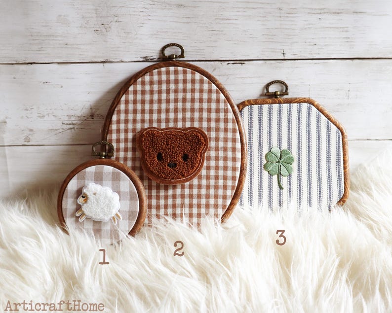 May include: Three embroidery hoops with fabric backgrounds and embroidered motifs. Hoop 1 is small and round with a white sheep. Hoop 2 is large and oval with a brown teddy bear. Hoop 3 is square with a green four-leaf clover. Text 1, 2, 3 and ArticraftHome.