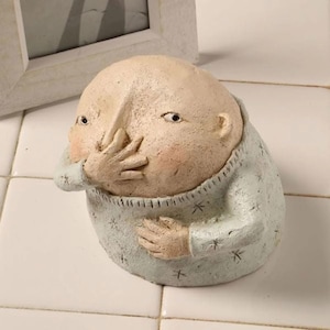 May include: A ceramic figurine of a person with a large head and a blue sweater. The person is covering their mouth with their hand.