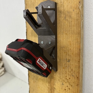 Puede incluir: Un soporte de pared de color marrón impreso en 3D para una batería de herramienta eléctrica roja y negra. La batería está etiquetada como "Milwaukee X20V TEAM".
