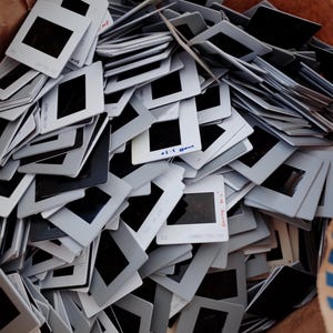 May include: A pile of vintage 35mm film slides. The slides have white frames with black rectangular windows. Some slides have handwritten text. The slides are in a brown paper bag.