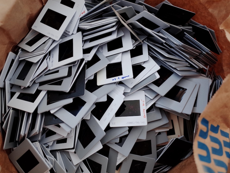 May include: A pile of vintage 35mm film slides. The slides have white frames with black rectangular windows. Some slides have handwritten text. The slides are in a brown paper bag.