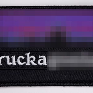 Op de afbeelding: Zwart rechthoekig embleem met een gepixeld zonsondergangontwerp in paars, blauw en zwart. Het woord "rucka" is in het wit geborduurd. Een wazige grijze rechthoek staat eronder.