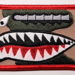 May include: Embroidered patch featuring a stylized, cartoonish shark face with a red border. The design includes a camouflage green upper section with gray teeth, a tan background, and a lower jaw with white, black, and red teeth.
