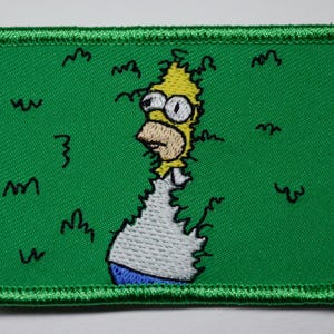 Könnte beinhalten: Gestickter Aufnäher mit Homer Simpson mit schockiertem Gesichtsausdruck. Der Aufnäher hat einen grünen Hintergrund mit schwarzen Kringeln und einen grünen Rand. Homers Haare sind gelb, seine Haut ist gebräunt und er trägt ein weißes Hemd und eine blaue Hose.