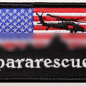 Puede incluir: Parche bordado con la bandera estadounidense, una silueta de helicóptero y la palabra "pararescue" en blanco. El parche tiene un borde negro y forma rectangular.
