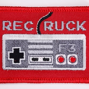 May include: Red embroidered patch featuring a gray and white controller design with the text "REC RUCK" above it. The controller has a directional pad, buttons, and the letters "F3". The patch has a red border.