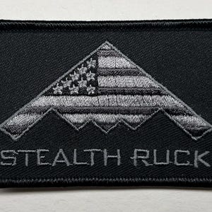May include: Black embroidered patch featuring a stylized B-2 stealth bomber design with an American flag motif. The text "STEALTH RUCK" is embroidered below the aircraft image. The patch has a black border.