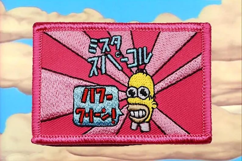 May include: A rectangular embroidered patch featuring a pink background with radiating dark pink sunburst lines. It depicts a yellow cartoon character head with wide eyes and a speech bubble containing Japanese text, plus Japanese text above.