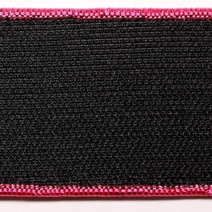 May include: A rectangular patch featuring a solid black center with a textured, woven fabric surface. The edges of the patch are finished with a thick, vibrant pink embroidered border that frames the black rectangular center completely.