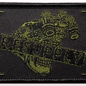 May include: Black rectangular patch featuring a green embroidered gas mask and the word "RESUPPLY". The patch has a black border and green corner details. The design is detailed and textured.