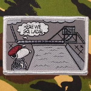 May include: Embroidered patch featuring Snoopy in a red beret, with the text "HERE WE GO, LADS!" in a thought bubble. The patch depicts a cartoon scene with a ramp and a structure, all in shades of gray and white, with a silver border.