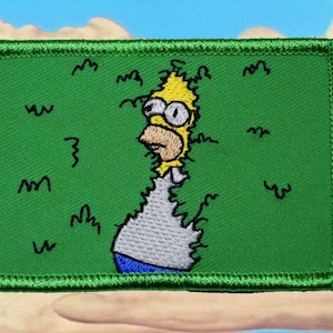 May include: Embroidered patch showing Homer Simpson breaking through a green background. The patch has a green border and depicts Homer with a shocked expression, yellow hair, and a white shirt. The background includes stylized grass.