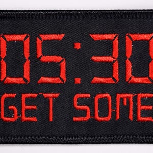 May include: Black rectangular patch with red embroidered text and numbers. The numbers "05:30" are above the words "GET SOME". The patch has a black border.