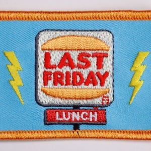 May include: Embroidered patch with a light blue background and an orange border. The patch features a burger graphic with the words "LAST FRIDAY" in red and "LUNCH" in red. Two yellow lightning bolts flank the burger.