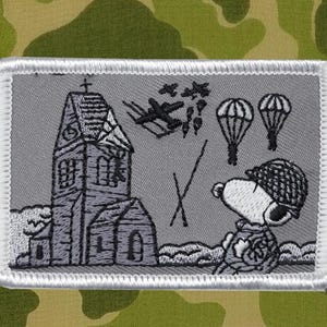 Könnte beinhalten: Gestickter Aufnäher mit Snoopy in einem Militärhelm, der eine Kirche, Flugzeuge und Fallschirme beobachtet. Der Aufnäher hat einen grauen Hintergrund mit weißem Rand, vor einem Tarnmuster.