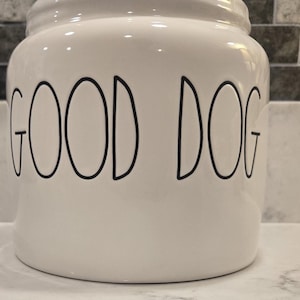 Puede incluir: Tarro de cerámica blanco con las palabras "GOOD DOG" en letras negras delineadas. El tarro tiene forma redondeada y tapa, ideal para guardar golosinas o comida para perros. El fondo presenta una encimera de tonos neutros y un salpicadero de azulejos decorativos.