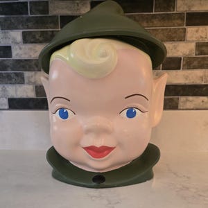 Puede incluir: Un tarro de galletas de cerámica con forma de cabeza de elfo con un sombrero verde. El elfo tiene el pelo rubio, los ojos azules y una cara sonriente.
