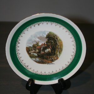 Puede incluir: Un plato de cerámica blanco con un borde verde y un ribete dorado. El plato presenta una ilustración vintage de una cabaña en un entorno rural con árboles y un arroyo. El texto "The Old Mill" está impreso en el plato.