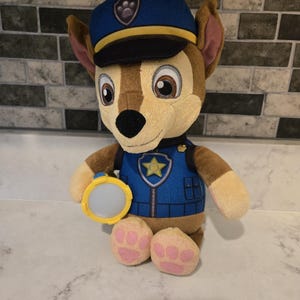 May include: A plush toy of a cartoon dog wearing a blue police hat and vest. The dog has brown fur, floppy ears, and pink paw pads. It holds a yellow flashlight. The vest has a star emblem.