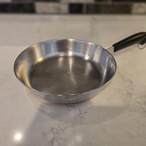May include: A stainless steel frying pan with a black handle. The pan has a shiny, reflective surface and a copper-coloured base. The handle has a loop for hanging. Kitchenware.