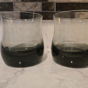 Op de afbeelding: Twee heldere glazen drinkbekers met een opvallend ombre-effect dat verloopt van een diep, donker antracietgrijs bij de afgeronde bodem naar een lichter, doorschijnend grijs bij de uitlopende rand. De glazen hebben een glad, gebogen silhouet.