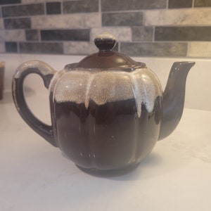 May include: A brown and white ceramic teapot with a rounded body and a handle. The teapot has a glazed finish with a white stripe around the top.