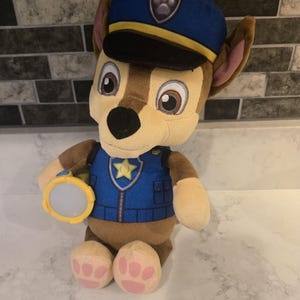 May include: A plush toy of Chase from Paw Patrol. The toy is brown and tan with a blue police hat and vest. Chase is holding a yellow flashlight. The toy has pink paw pads and large, expressive eyes.