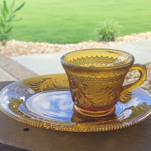 Puede incluir: Un juego de tazas de té y platillo de vidrio ámbar con un delicado patrón floral. Las tazas y el platillo tienen un aspecto vintage y son perfectas para una merienda.