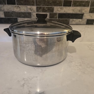 May include: A round, silver-coloured metal cooking pot with a matching lid. The pot features two black plastic side handles and a black plastic knob on the centre of the lid. The exterior surface of the metal pot has a slightly weathered finish.