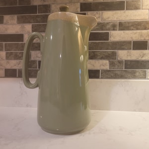 以下が含まれることがあります： クリーム色の蓋と注ぎ口が付いた、背の高いセージグリーンのセラミックコーヒーポット。ポットには大きなハンドルと丸みを帯びたベースが付いています。背景にはニュートラルなトーンのレンガ模様があります。