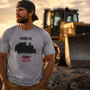 May include: A heather grey short-sleeve t-shirt featuring a black silhouette of a bulldozer. The text reads LEARN TO, followed by the word CODE with a red line through it, and the word WELD in red text below the crossed-out word.