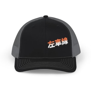 Puede incluir: Gorra de camionero negra con parte trasera de malla gris y visera curva. El panel frontal muestra los caracteres japoneses "左車線" en blanco y naranja. Cierre trasero ajustable.