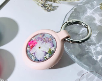 May include: A pink keychain with a clear round charm containing pressed flowers. The charm is attached to a silver metal ring and a silver metal clip.