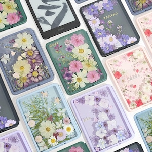 May include: A collection of rectangular e-reader cases featuring clear shells filled with various arrangements of pressed dried flowers in shades of pink, purple, white, and green, with some cases displaying custom names in gold script lettering.