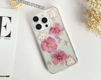 May include: A clear phone case with dried pink and white flowers pressed inside. The case is designed for an iPhone with a triple lens camera system.