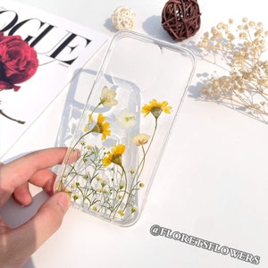May include: A clear phone case with pressed yellow and white flowers. The case has a floral design with small white flowers and larger yellow flowers with a yellow center. The case is on a white surface with a magazine and dried flowers in the background. The text "@floretsflowers" is visible in the bottom right corner of the image.