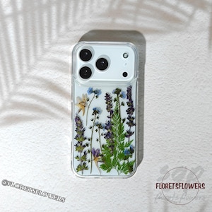 Puede incluir: Funda transparente para teléfono con flores prensadas, incluyendo flores azules, moradas y amarillas. La funda tiene una parte superior blanca con lentes de cámara y un flash. El texto "@FLORETSFLOWERS" es visible abajo a la izquierda.