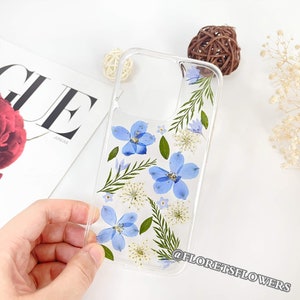 Op de afbeelding: Een transparante telefoonhoes met geperste blauwe en witte bloemen en groene bladeren. De hoes heeft een transparante achterkant en zijkanten, en de bloemen zijn zichtbaar door de hoes. De hoes is ontworpen om te passen op een smartphone. De tekst "@floretsflowers" is afgedrukt in de rechterbenedenhoek van de hoes.