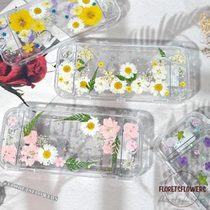 Op de afbeelding: Transparante plastic hoesjes versierd met geperste bloemen, waaronder madeliefjes, gele bloemen en roze bloemen. De hoesjes zijn voor een game-apparaat. De hoesjes hebben de tekst "FLORETSFLOWERS" en "@FLORETSFLOWERS".